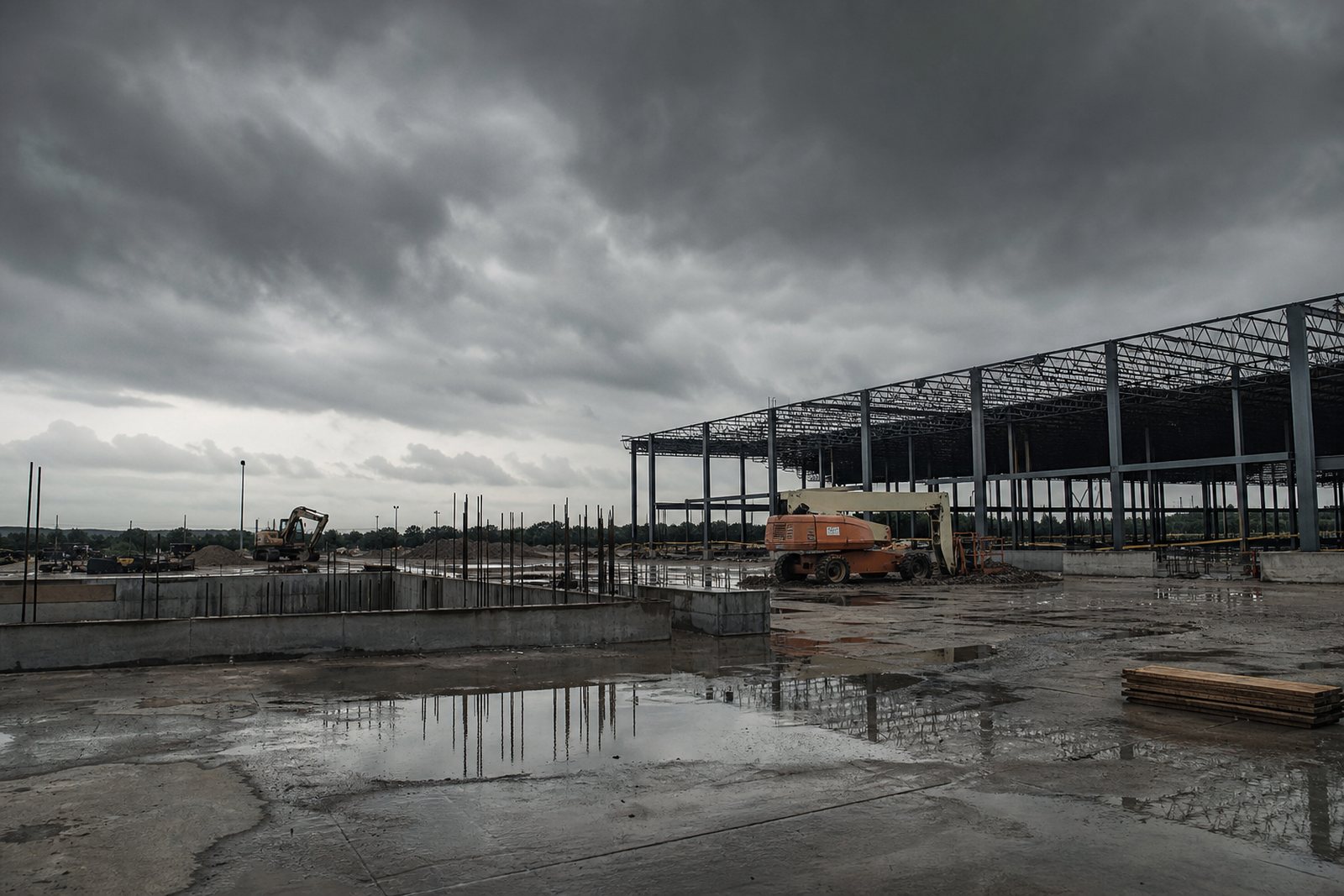 Commercial construction site under heavy grey clouds with standing water on poured concrete and idle equipment